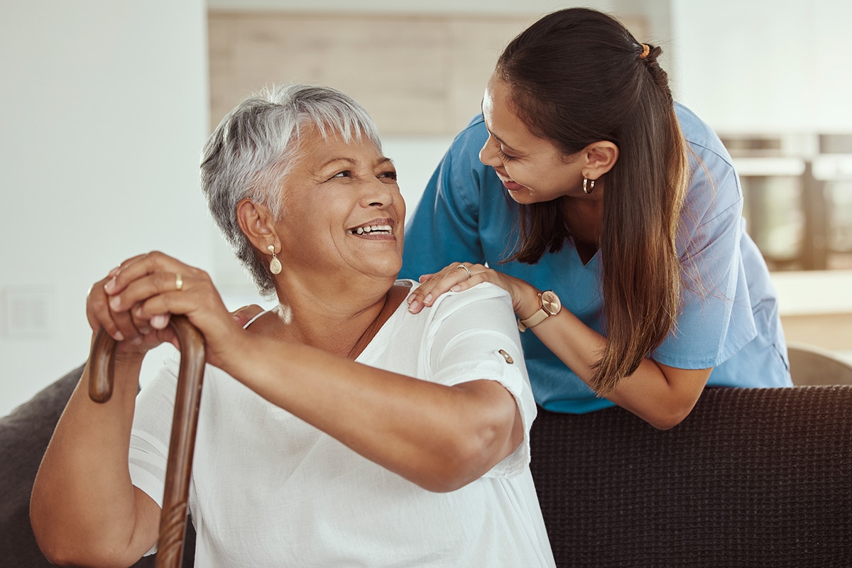 Happy, relax and senior woman with caregiver smile while sitting on a living room sofa in a nursing home. Support, help and professional nurse or healthcare worker helping elderly lady or patient